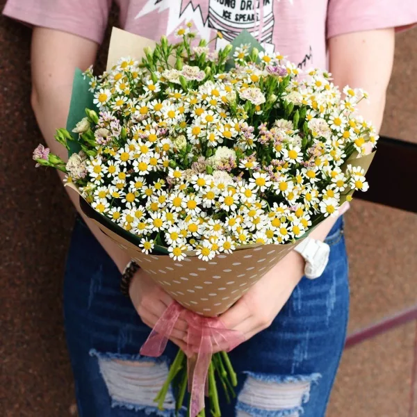 Пышный букет из белого танацетума с жёлтыми сердцевинами в крафт-упаковке с розовой лентой