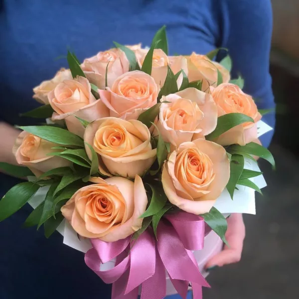 Коробка из 13 персиково-кремовых роз куполом с атласной розовой лентой, в руках