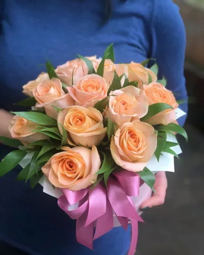 Коробка из 13 персиково-кремовых роз куполом с атласной розовой лентой, в руках
