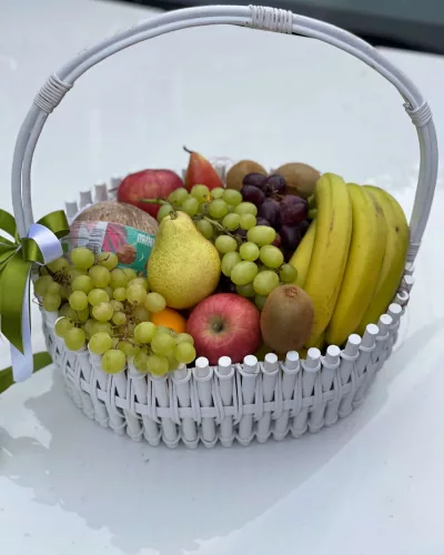 Белая плетёная корзина с жёлтыми бананами, зелёным и чёрным виноградом, грушей и красными яблоками, декоративные бусы